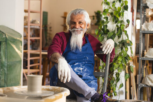 Happy biracial senior potter with long beard sitting next to potter's wheel in pottery studio