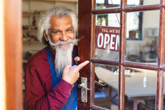 Happy biracial senior business owner with long beard showing shop sign to open