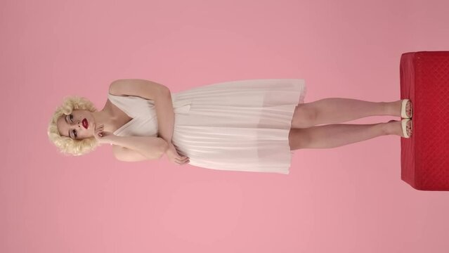Woman in the look of Marilyn Monroe, dreamily thinking about something, putting her fingers to her lips, there is an idea. Woman standing full length on a pedestal on a pink. Vertical video.