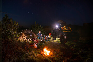 Night starry sky. car and hikers near campfire. Couple man and woman sitting near bonfire under majestic blue sky with stars. camping, travelling tourist things, tent chairs, table.