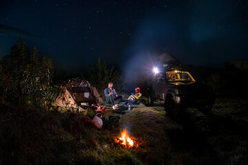 They talk and drink. car and hikers near campfire. Couple man and woman sitting near bonfire under majestic blue sky with stars. camping, travelling tourist things, tent chairs, table.