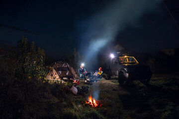 Night starry sky. Smoke and headlamp light. Vehicle near campfire. Couple man and woman sitting near bonfire under majestic blue sky with stars. camping, travelling tourist things, tent chairs, table. - Powered by Adobe