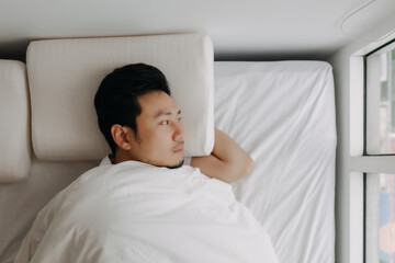 Asian man lying alone on white bed with soft pillow and blanket, thinking of something, looking at...