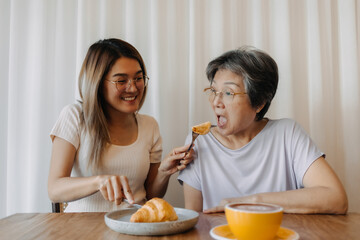 Obraz na płótnie Canvas Asian old woman mother and adult daughter smiling, one giving and feeding mom eating croissant bakery, spending time together in cafe, happy good life.