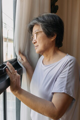 Asian old woman opening curtain while looking out the window, missing and waiting family and thinking something alone.