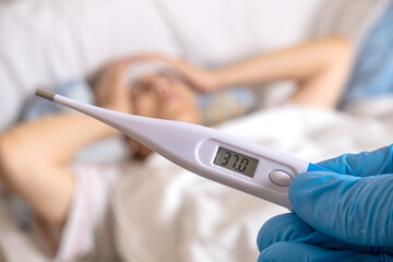 Doctor checking temperature of senior woman lying in bed with digital thermometer