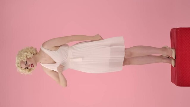 A woman shouting, swearing, accusing someone, pointing finger at him. Woman in look like Marilyn Monroe exhales, fixes her hair, smiles. Woman standing full length on pedestal on pink. Vertical video.
