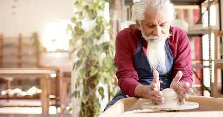 Focused senior biracial potter with long beard using potter's wheel in pottery studio, slow motion