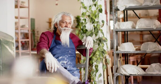 Happy senior biracial potter with long beard resting and smiling in pottery studio, slow motion