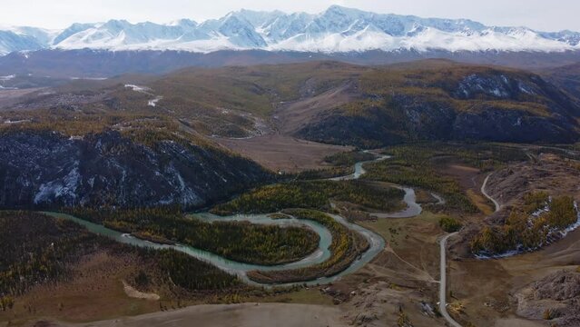 Slow motion aerial view of green tree hills with meandering flowing river water and motorable roadway in Altai, Russia against snow capped mountains cloudless blue sky in daylight
