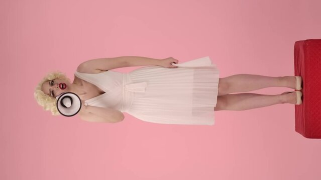 Woman in character of Marilyn Monroe speaking into megaphone. Spectacular woman standing on red pedestal full-length in studio on pink background. Vertical video. Sale, Black Friday. Copy space.