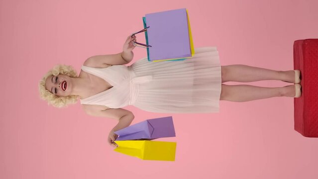 Young happy woman holding colorful shopping bags. Woman in the image of Marilyn Monroe standing full length on pedestal in studio on pink background. Vertical video. Shopping concept, sales.