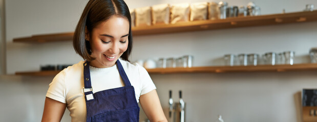 Vertical shot of smiling asian girl putting coffee in tamper to brew capuccino, prepare order in...