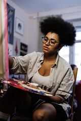 African american artist holding paint palette near blurred canvas in workshop 