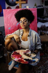 African american artist holding palette and paintbrush near canvas on easel in workshop