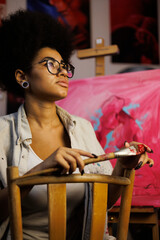 African american artist holding paintbrush and looking away near blurred easel and canvas in art studio 