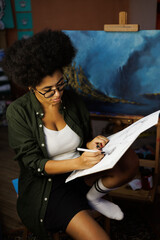 African american female artist drawing on sketchpad near painting in studio in evening