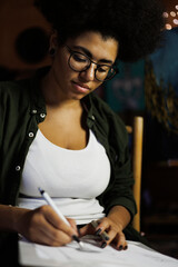 Obraz na płótnie Canvas Portrait of african american artist in eyeglasses drawing on sketchbook in studio with lighting 
