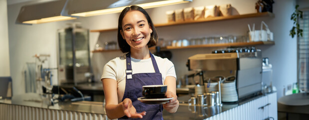 Portrait of smiling asian woman holds cup of coffee, prepare drinks for clients in cafe, working...