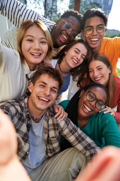 Happy group of multiethnic classmate taking a selfie together outdoors in the university campus, looking at camera cheerfully raising arms, celebrating last day. Vertical photo.