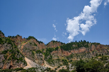 A view of a big mountain and tall rocks covered with trees and a blue cloudy sky