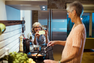 Senior lesbian couple capturing a cooking moment on a smartphone