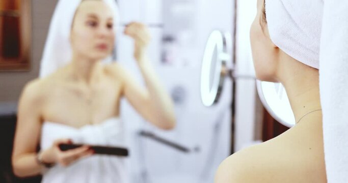 Young Woman Applying Make Up. SLOW MOTION. Girl apply nude eye shadow with brush, standing before the mirror in the bathroom after shower. Everyday beauty rituals.