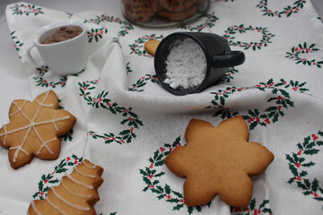 Christmas cookies, snowflakes or tree. Small white cup with cocoa powder and a fallen black cup spills powdered sugar. White tablecloth with green Christmas wreaths. A glass jar with chocolate cookies