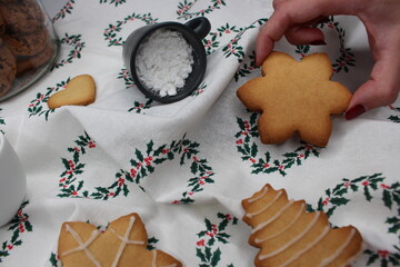 Female hand takes a snowflake cookie. In the background, a white tablecloth with Christmas wreaths, Christmas cookies, a black small cup dropping icing sugar and a a glass jar with chocolate sweets.