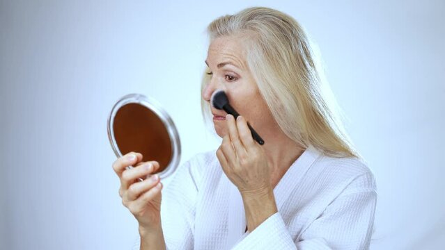 Looking in a mirror a mature middle aged woman applies natural makeup powder foundation with cosmetic brush on clean face skin smoothing wrinkles old lady looking in mirror beauty, white background