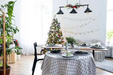 Festive interior of house is decorated for Christmas and New Year in loft style with black stove, fireplace, Christmas tree. Warm studio room with set table, burning wood, cozy and heating of home