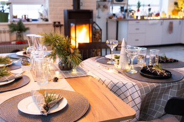 Serving festive table with plates, forks, knives, napkins, glasses close-up near stove fireplace in interior of loft house decorated for Christmas and New Year. Waiting for guests for festive dinner