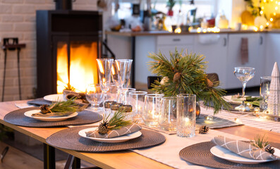 Serving festive table with plates, forks, knives, napkins, glasses close-up near stove fireplace in interior of loft house decorated for Christmas and New Year. Waiting for guests for festive dinner