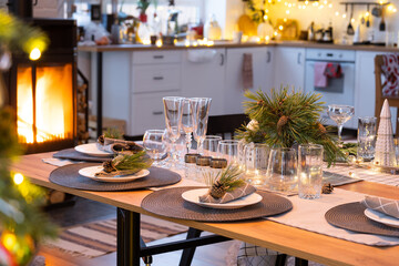 Serving festive table with plates, forks, knives, napkins, glasses close-up near stove fireplace in interior of loft house decorated for Christmas and New Year. Waiting for guests for festive dinner