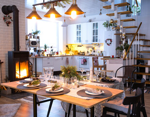 Festive interior of house is decorated for Christmas and New Year in loft style with black stove, fireplace, Christmas tree. Warm studio room with set table, burning wood, cozy and heating of home