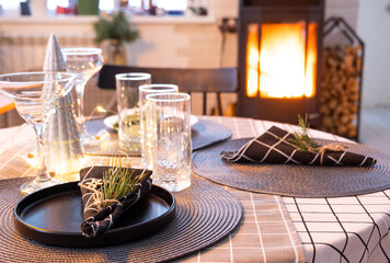 Serving festive table with plates, forks, knives, napkins, glasses close-up near stove fireplace in interior of loft house decorated for Christmas and New Year. Waiting for guests for festive dinner