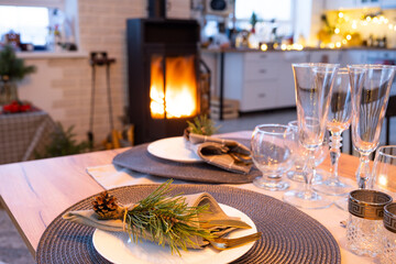 Serving festive table with plates, forks, knives, napkins, glasses close-up near stove fireplace in interior of loft house decorated for Christmas and New Year. Waiting for guests for festive dinner