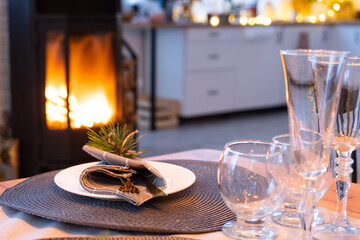Serving festive table with plates, forks, knives, napkins, glasses close-up near stove fireplace in interior of loft house decorated for Christmas and New Year. Waiting for guests for festive dinner