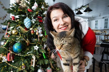 Selfie funny portrait of a woman in a red Santa dress and domestic cat in a home interior with a Christmas tree and New Year decor. Preparation for the holidays, party
