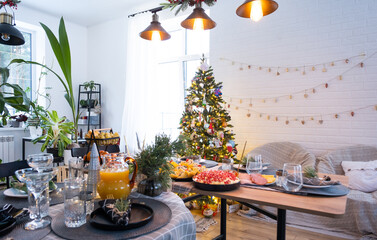 Festive interior of house is decorated for Christmas and New Year in loft style with black stove, fireplace, Christmas tree. Warm studio room with set table, burning wood, cozy and heating of home
