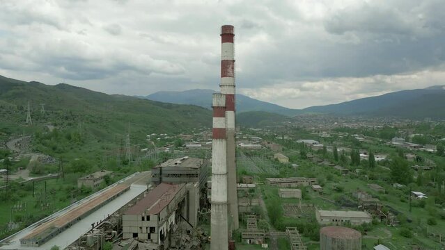 An industrial zone, an old ruined factory, chimneys, buildings with broken windows and trees on the roofs. Around high green mountains in cloudy weather. Drone video. Climbing along the pipes.