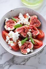 Bowl of fig fruit, tomato and goat cheese salad, vertical shot on a white marble background, close-up