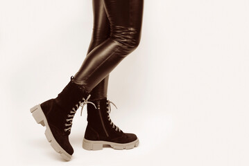A woman in leather black pants tries on high black suede boots with a high aggressive sole on a light background, chooses practical warm shoes for every day - obrazy, fototapety, plakaty