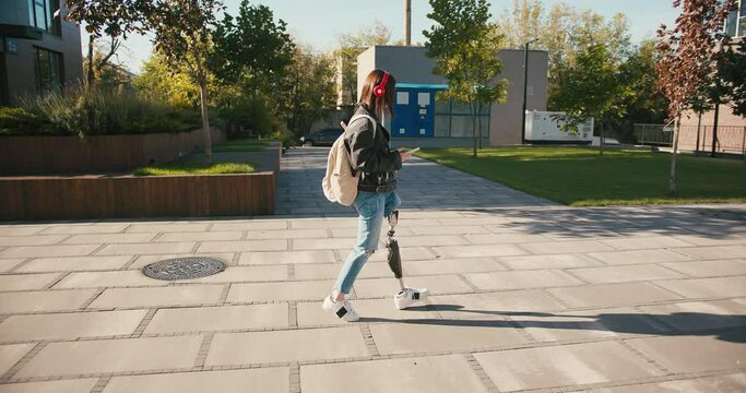 Young woman student with prosthetic leg walking in university campus. Woman with bionic leg. Woman with leg prosthesis equipment and smartphone