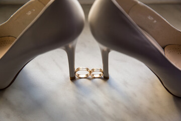 Elegant beige heels beside gold wedding rings, lit by soft natural light. Top-down shot capturing...