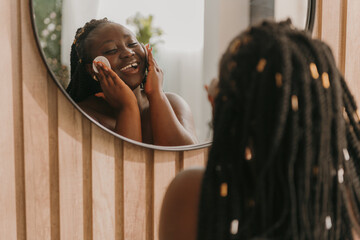 Happy plus size African woman cleaning face with cotton pad and looking at the mirror in bathroom - obrazy, fototapety, plakaty