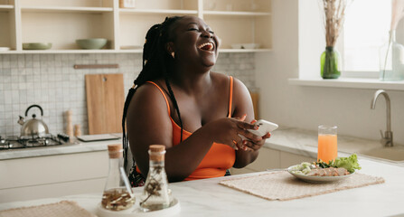 Beautiful plus size African woman using smart phone and laughing while enjoying healthy lunch at home