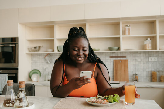 Beautiful plus size African woman using smart phone while enjoying healthy eating for lunch at home