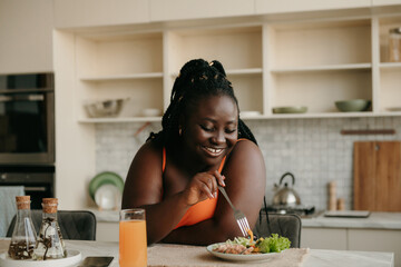 Beautiful curvy African woman enjoying healthy eating for lunch at home - obrazy, fototapety, plakaty