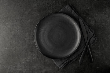 Empty grunge dark grey craftmade ceramic plate with cutlery and linen serviette on black stone...